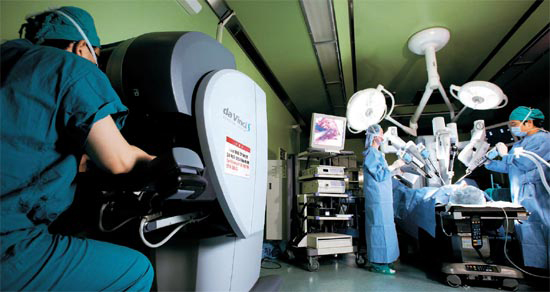 執刀医（左）が患者から離れたコンソールボックスで操縦桿を操作すればロボットアームが動き、手術を行う。