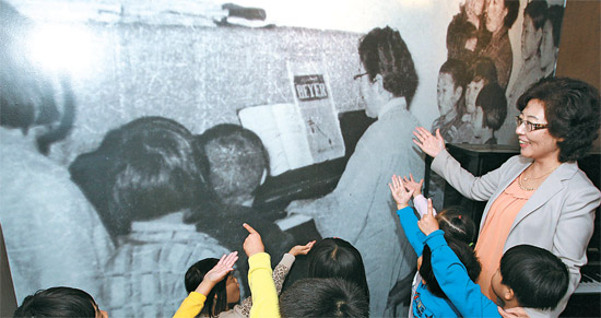 尹鶴子のピアノ演奏の姿を拡大したパネルの前で孫娘のチョン・エラ院長（右）が子どもたちに話している。