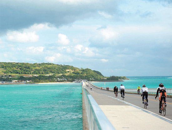 日本の沖縄県はスポーツを観光産業の活性化の素にするために努力している。一年中開かれるマラソン大会は約３０回に達する。写真は沖縄本島と古宇利島をつなぐ古宇利大橋でサイクリングを楽しむ観光客。エメラルド色の海を満喫できる。（写真＝沖縄スポーツコンベンションビューロー）