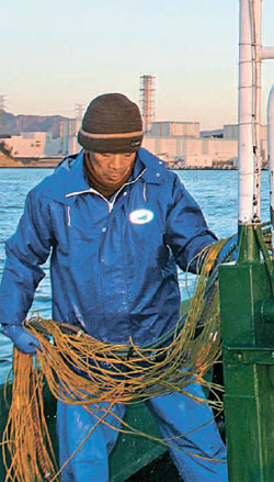 漁船の船員の泉さんが海に投げた網を巻き上げている。後ろには福島第１・２原発より南側にある火力発電所が見える。