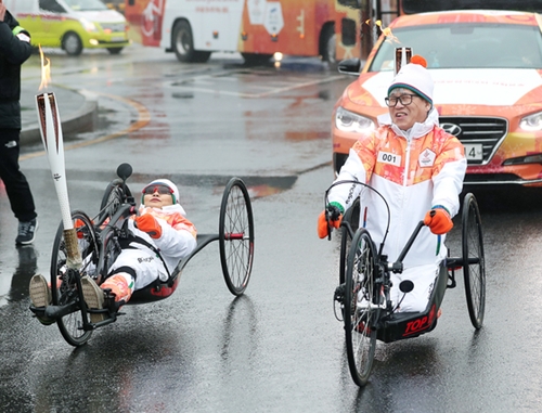 ４日、２０１８年平昌冬季パラリンピックのソウル区間聖火リレーがソウル蚕室総合運動場からスタートし、第一走者として登場したソウル市障害者サイクル連盟のナ・ウンファさん（左）、チェ・ムンギさん（右）が車椅子サイクルに乗って聖火を運んでいる。